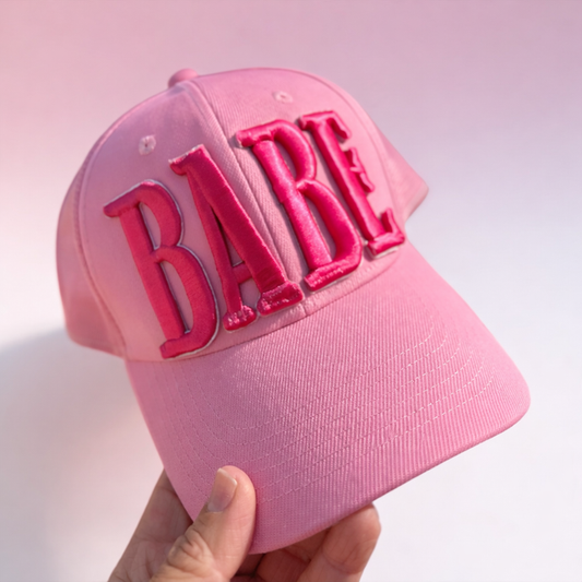 Pink cap with 'Babe' text held by a hand against a light pink background