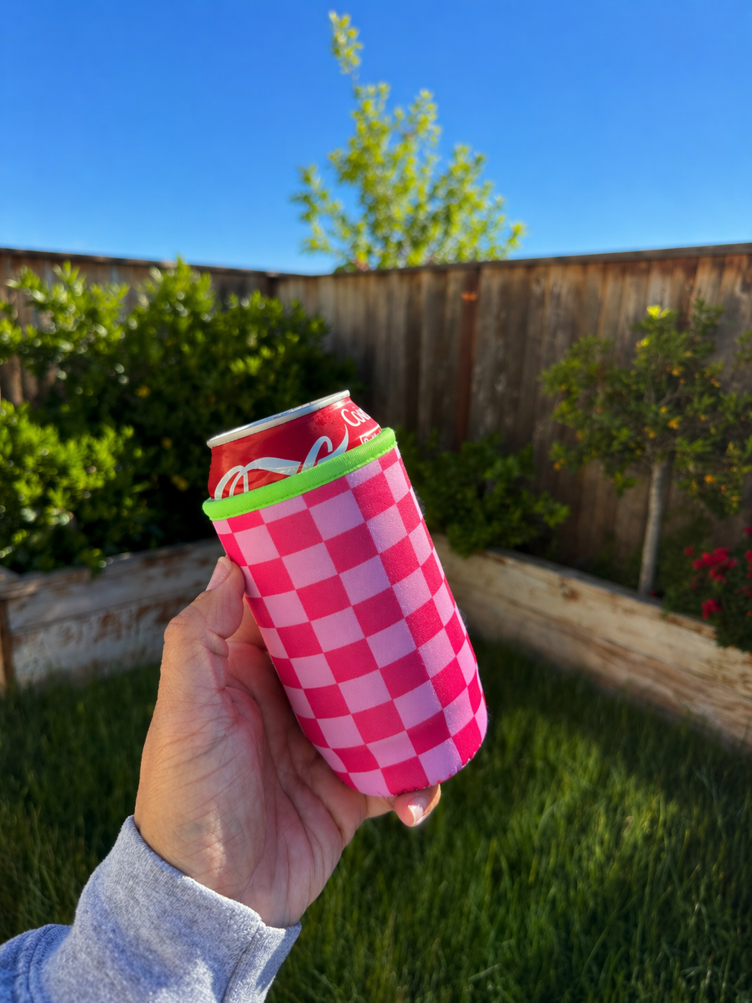 Hand holding a can with a pink checkered sleeve against a garden background